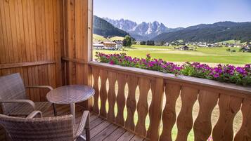 A wooden balcony with wicker chairs and a small table, overlooking a green valley and mountains.