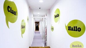 A hallway with a carpeted floor and white walls. There are several speech bubbles with the word "hola" in different languages.