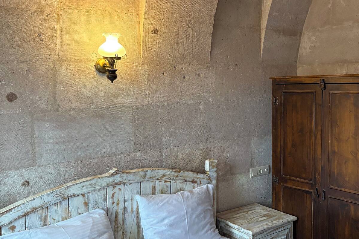 Una habitació amb un llit, una tauleta de nit, una làmpada penjada a la paret i un armari de fusta.