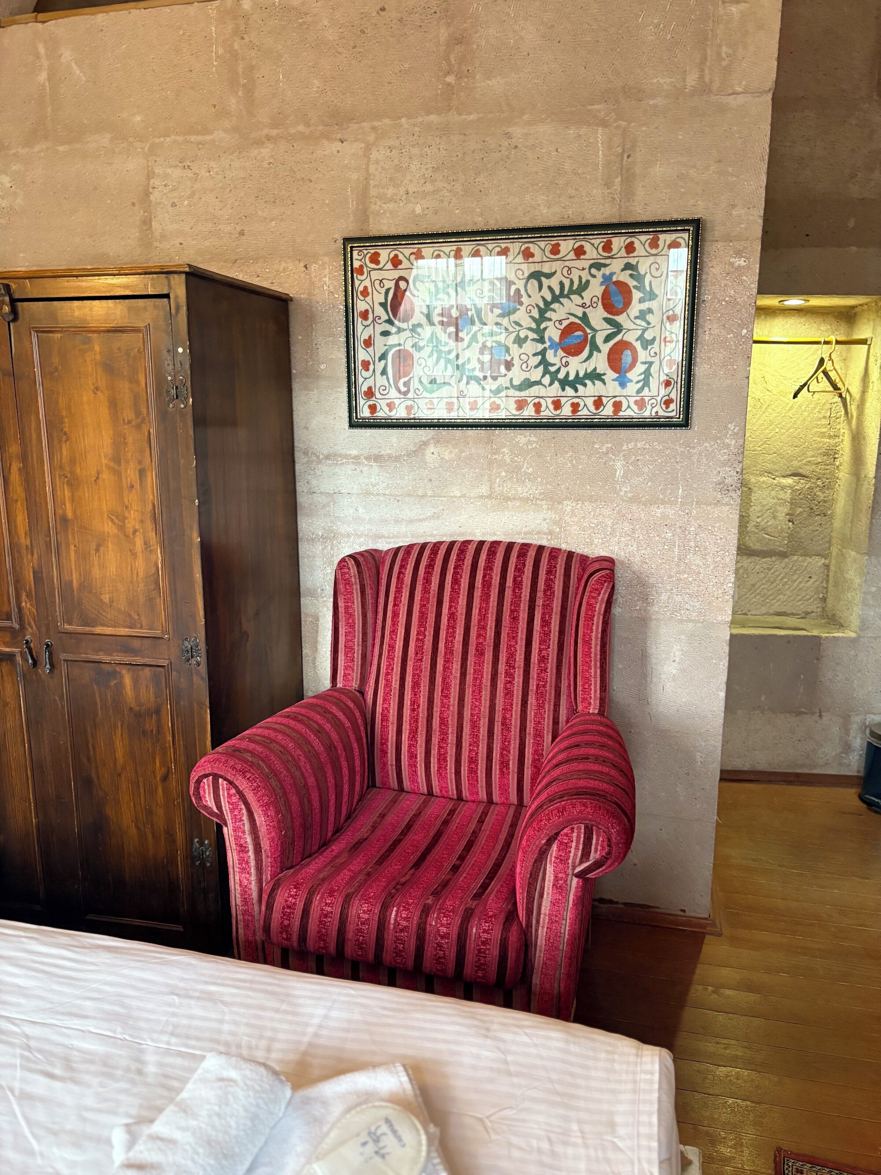 Una habitación con un armario de madera, un sillón rojo a rayas, un adorno en la pared y una cama con colcha blanca.