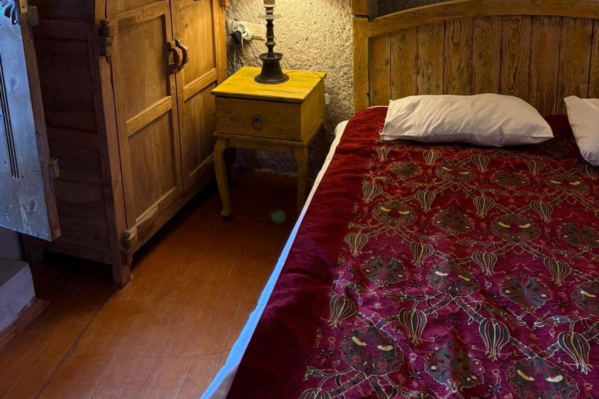 Un dormitori d'àtic acollidor amb un armari de fusta, una taula de nit amb una làmpada i un llit amb un llençol de llit vermell amb dibuixos.