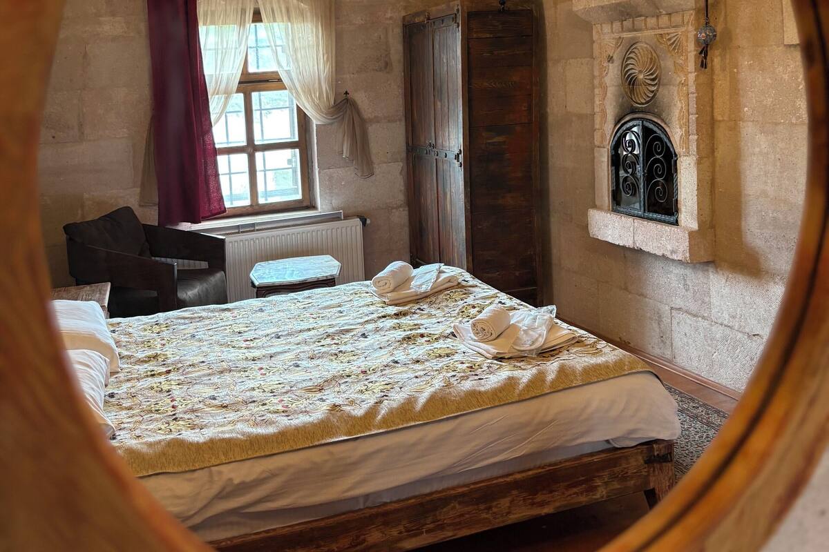 Habitació amb llit, finestra amb cortines, radiador, porta de fusta i paret de pedra.