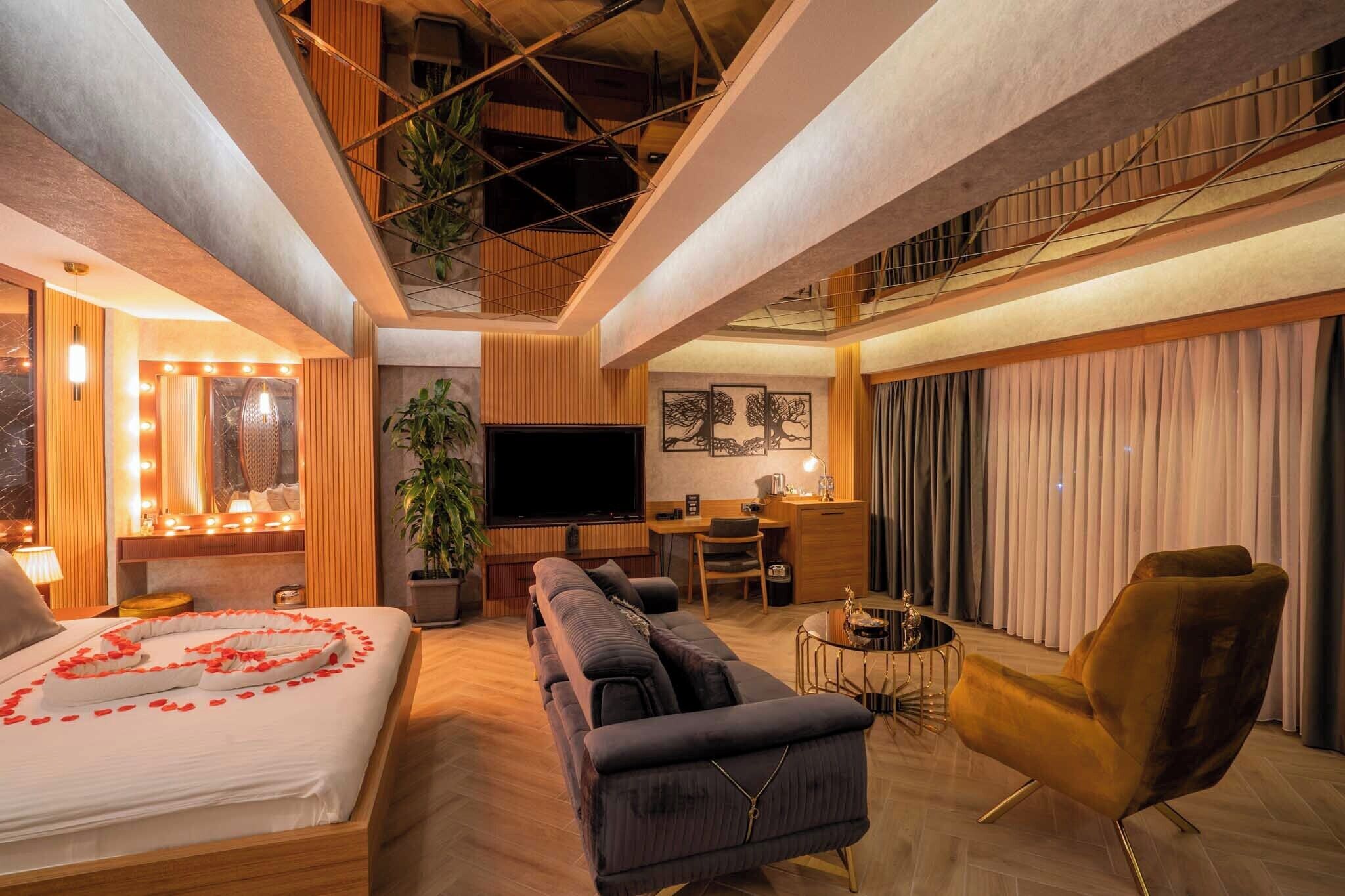 A modern hotel room with a bed, sofa, armchair, desk, and a large TV.