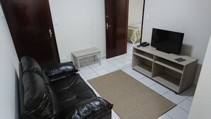 A living room with a black leather sofa, a small white bench, a television on a wooden stand, and a tiled floor.