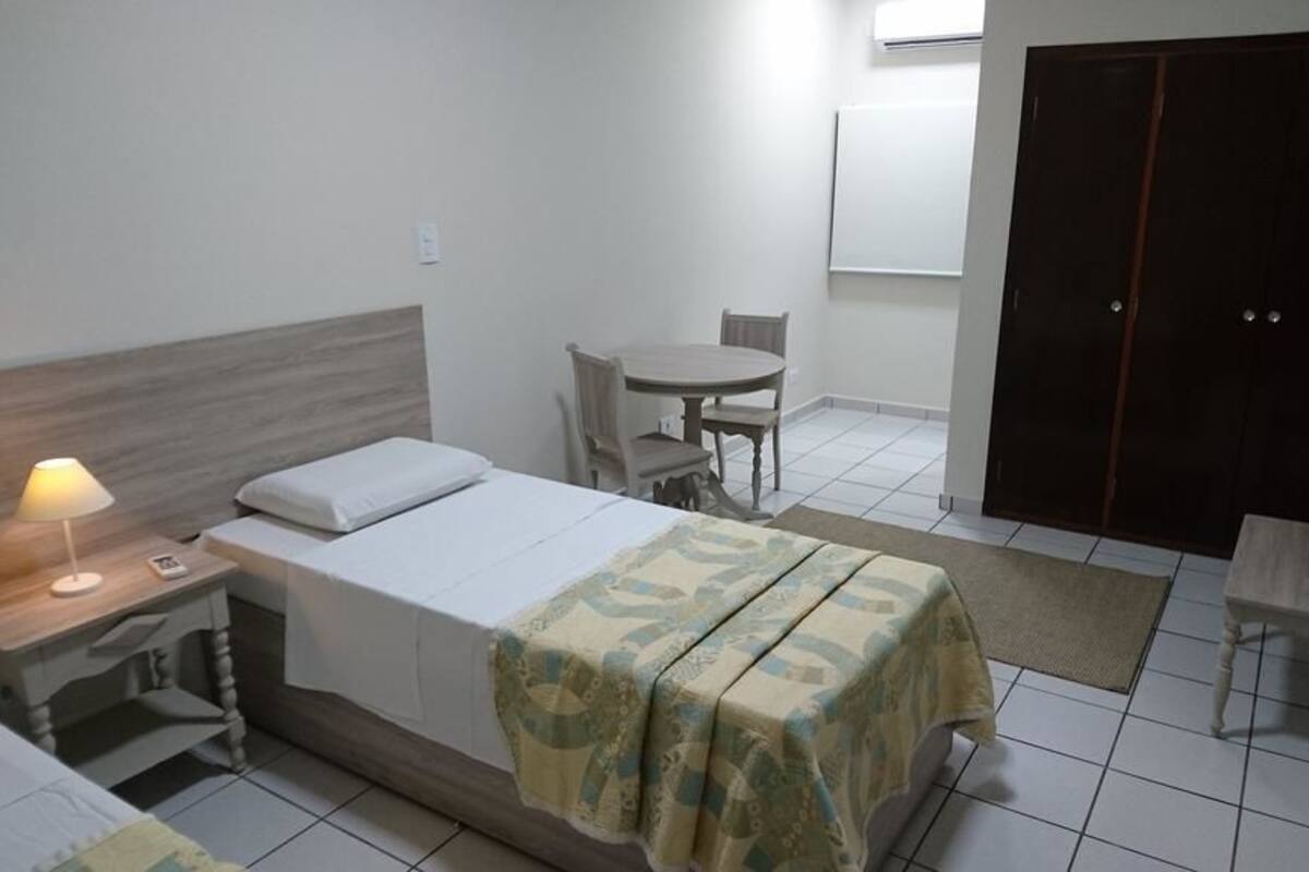 Un lit simple avec une couvre-lit à motifs, une table de chevet avec une lampe, une petite chaise, un bureau et une armoire.