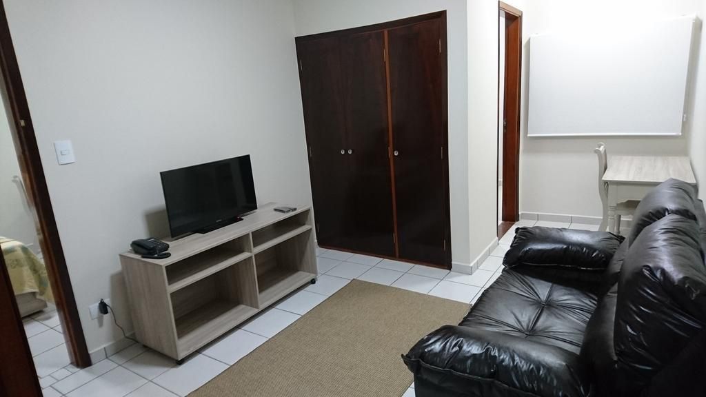 Uma sala de estar com televisão sobre um suporte de madeira, um sofá de couro preto, um guarda-roupa e uma mesinha de cabeceira com abajur.