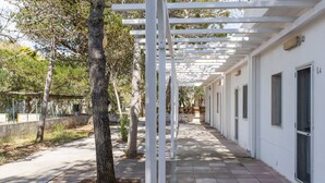 A covered walkway with white columns, trees, and a pathway leading to a row of white buildings with windows. - Centro Vacanze Lido Smeraldo (Lecce)