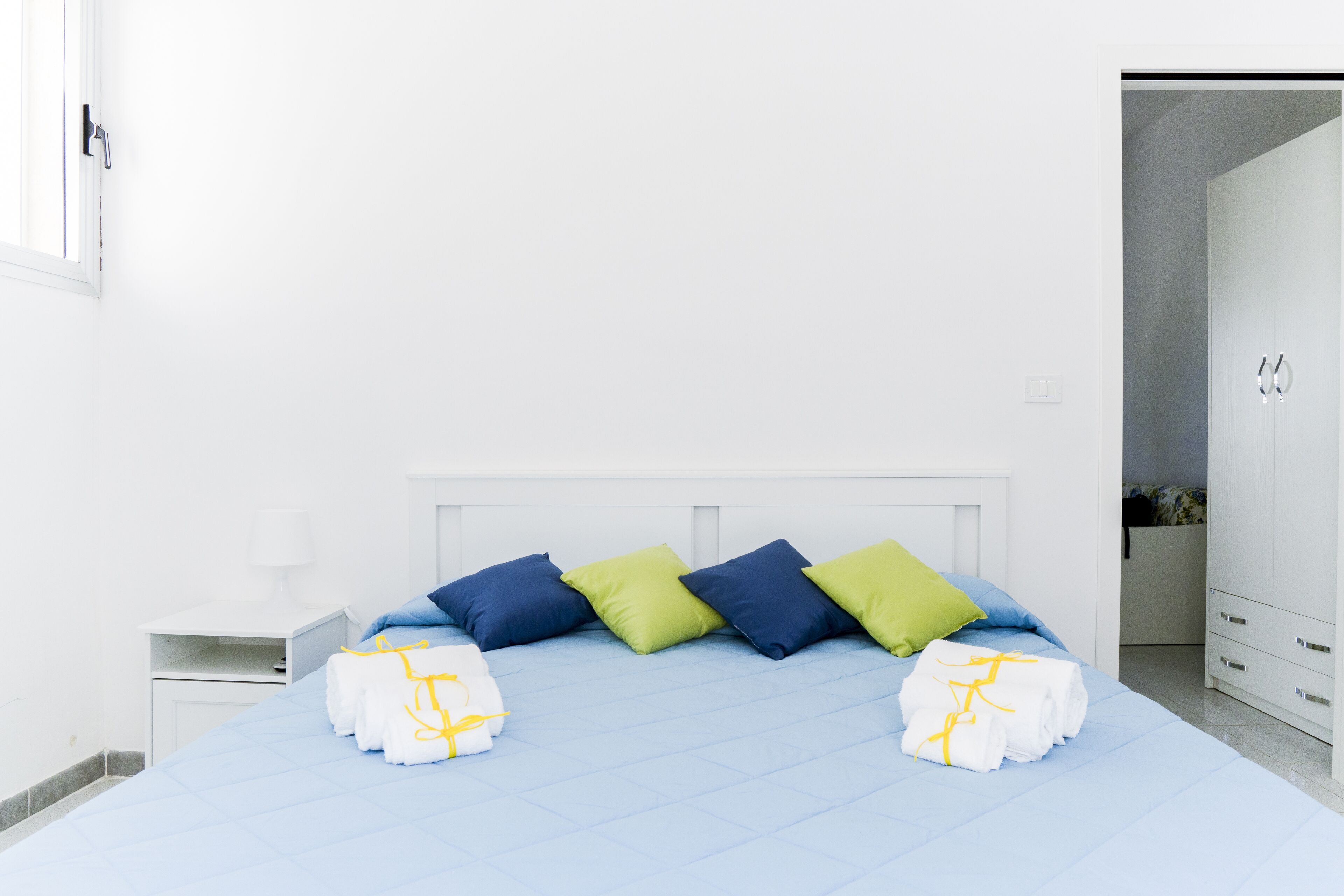 Un lit bien fait, agrémenté d’oreillers bleus et verts, une table de chevet blanche avec une lampe, et une armoire blanche.