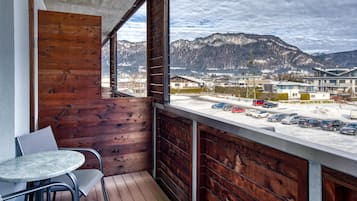 A balcony with a wooden wall, a round table, and chairs. It overlooks a parking area and buildings with mountains in the background.