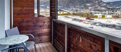 A balcony with a wooden wall, a round table, and chairs. It overlooks a parking area and buildings with mountains in the background.