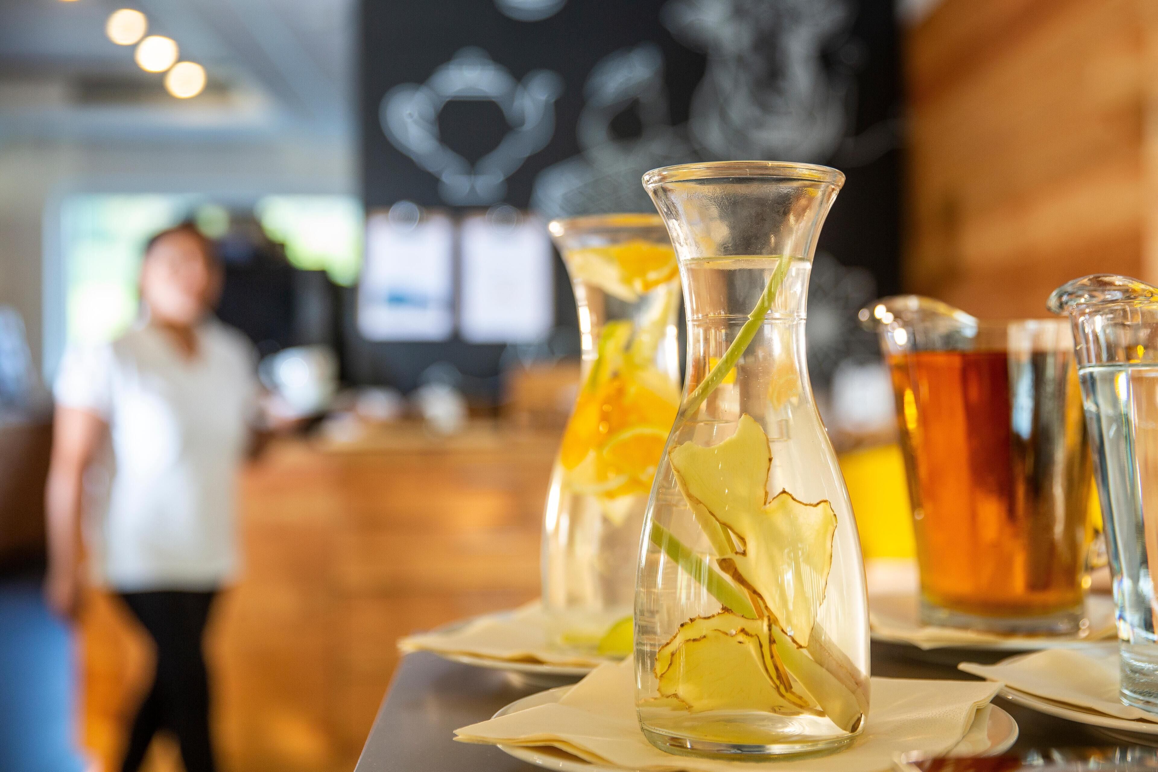 A bar area with clear glass vases containing beverages and garnishes, a person in the background, and a chalkboard with writing.