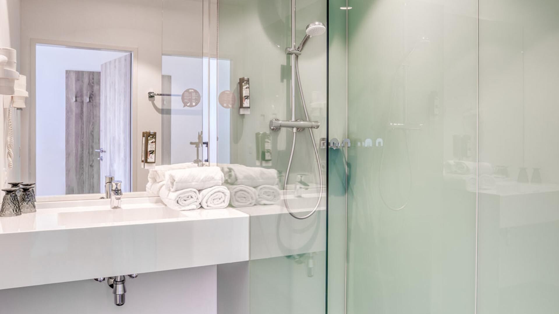 Een moderne badkamer met een glazen douchecabine, een witte wastafel met een stap handdoeken en een grote spiegel.