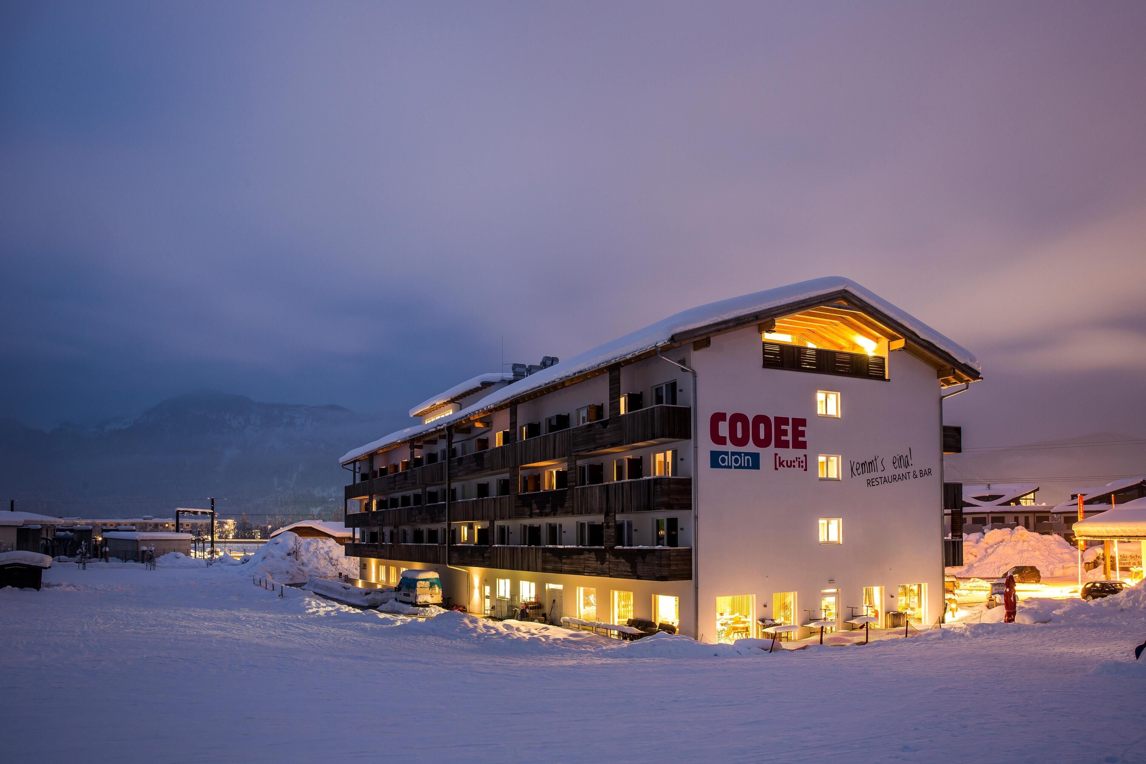 Ein mehrstöckiges Hotel mit verschneitem Dach, beleuchteten Fenstern und einem Schild mit der Aufschrift "COOEE alpin".
