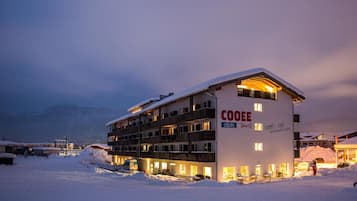 A multi-story hotel with a snow-covered roof, illuminated windows, and a sign reading "COOEE alpin".