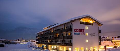 Ein mehrstöckiges Hotel mit verschneitem Dach, beleuchteten Fenstern und einem Schild mit der Aufschrift "COOEE alpin".