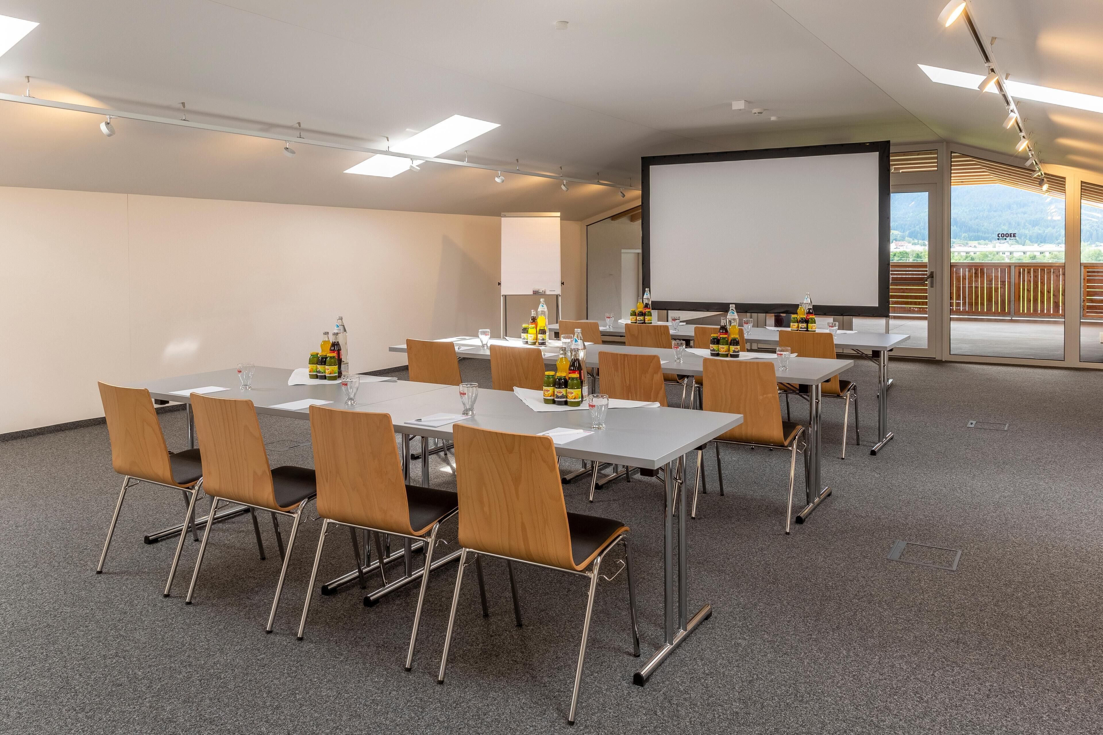 A conference room with tables, chairs, a projector screen, and a view of a balcony.