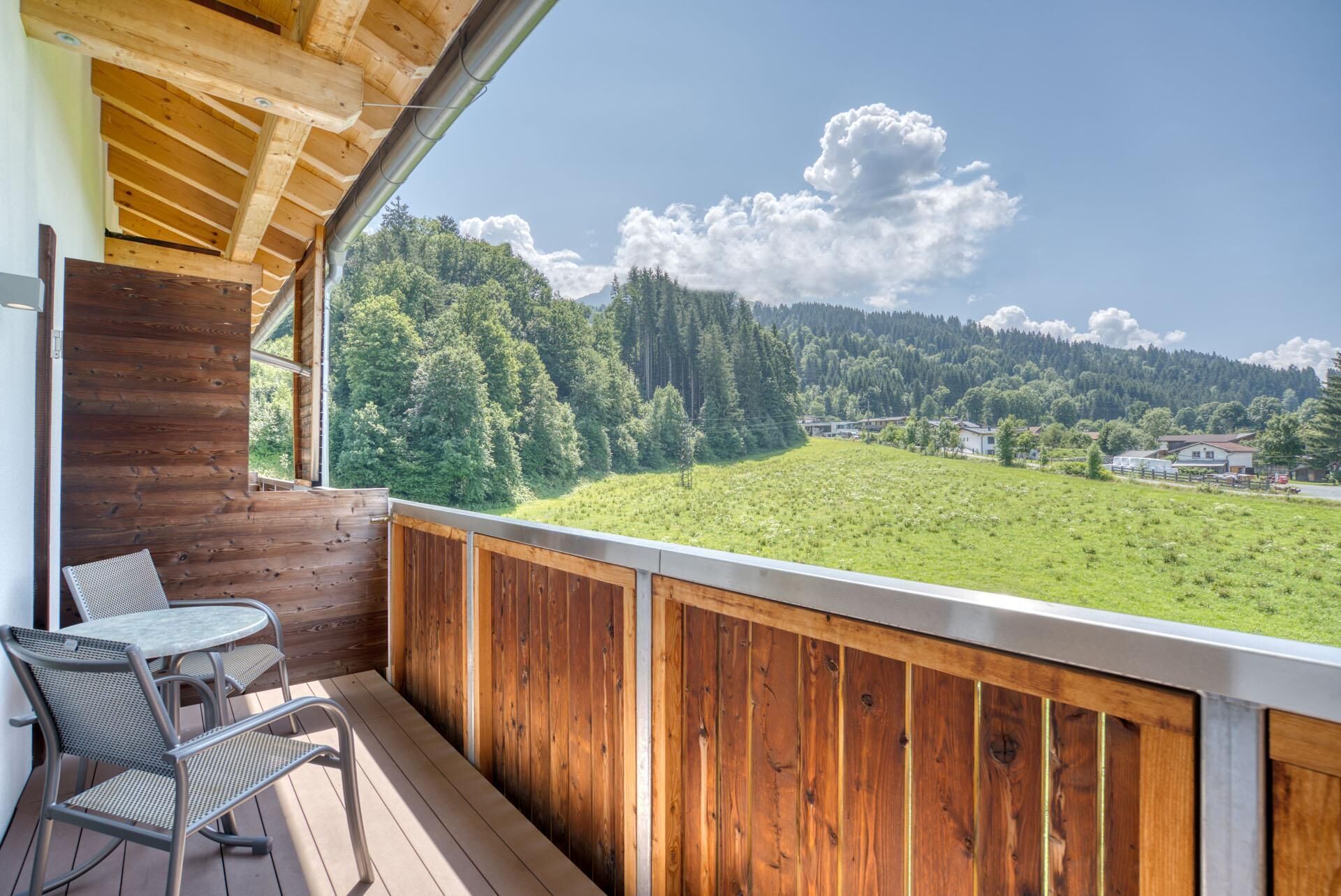 A wooden balcony with a view of a green field and forested hills.