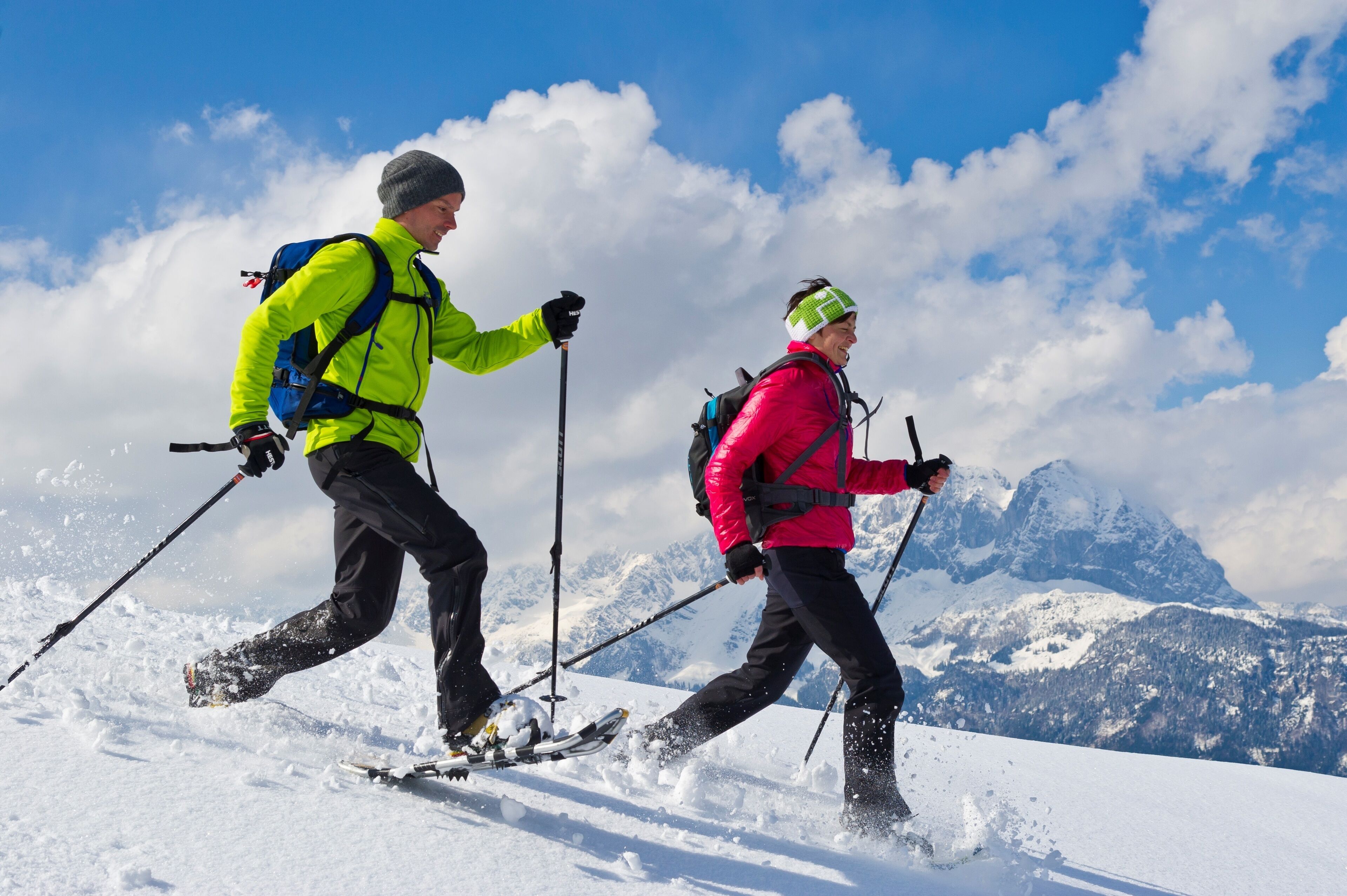 Two people skiing on a snowy mountain with a clear sky and snow-covered peaks in the background.