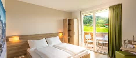 A double bed with a wooden headboard, a nightstand with a lamp, a green bench, and a view of a patio with outdoor seating.