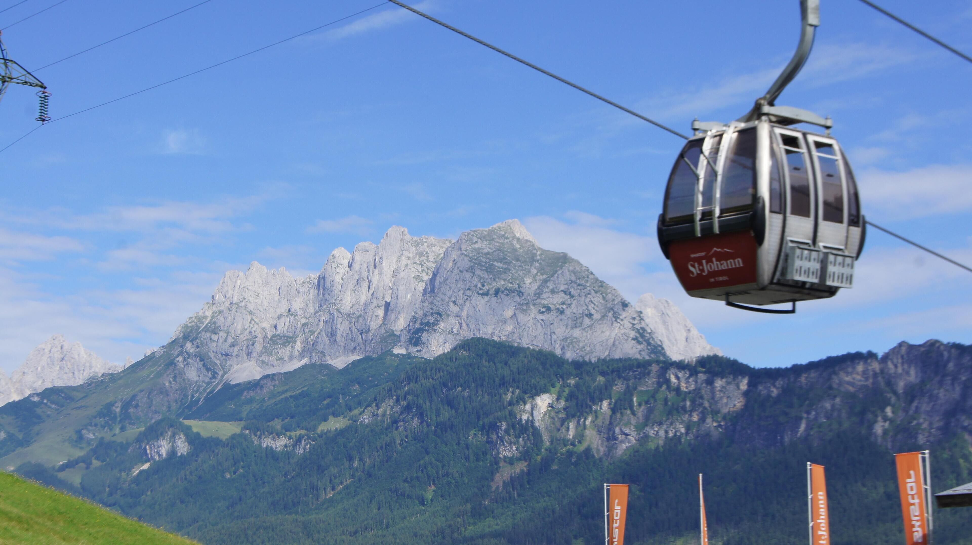 A cable car with "St. Johann" branding, a mountainous backdrop, and flags with "St. Johann" text.