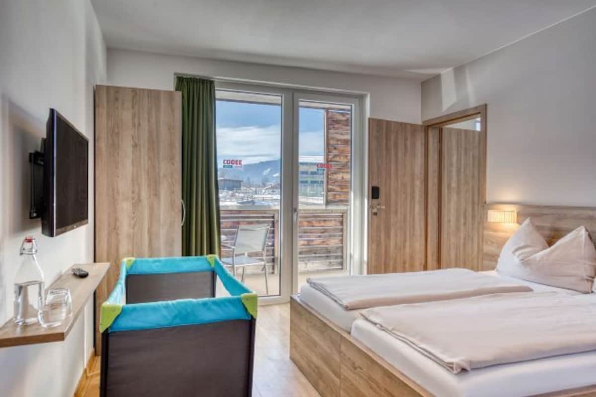 A hotel room with a bed, a small table with a bottle and glasses, a TV, and a view of a cityscape through a large window.