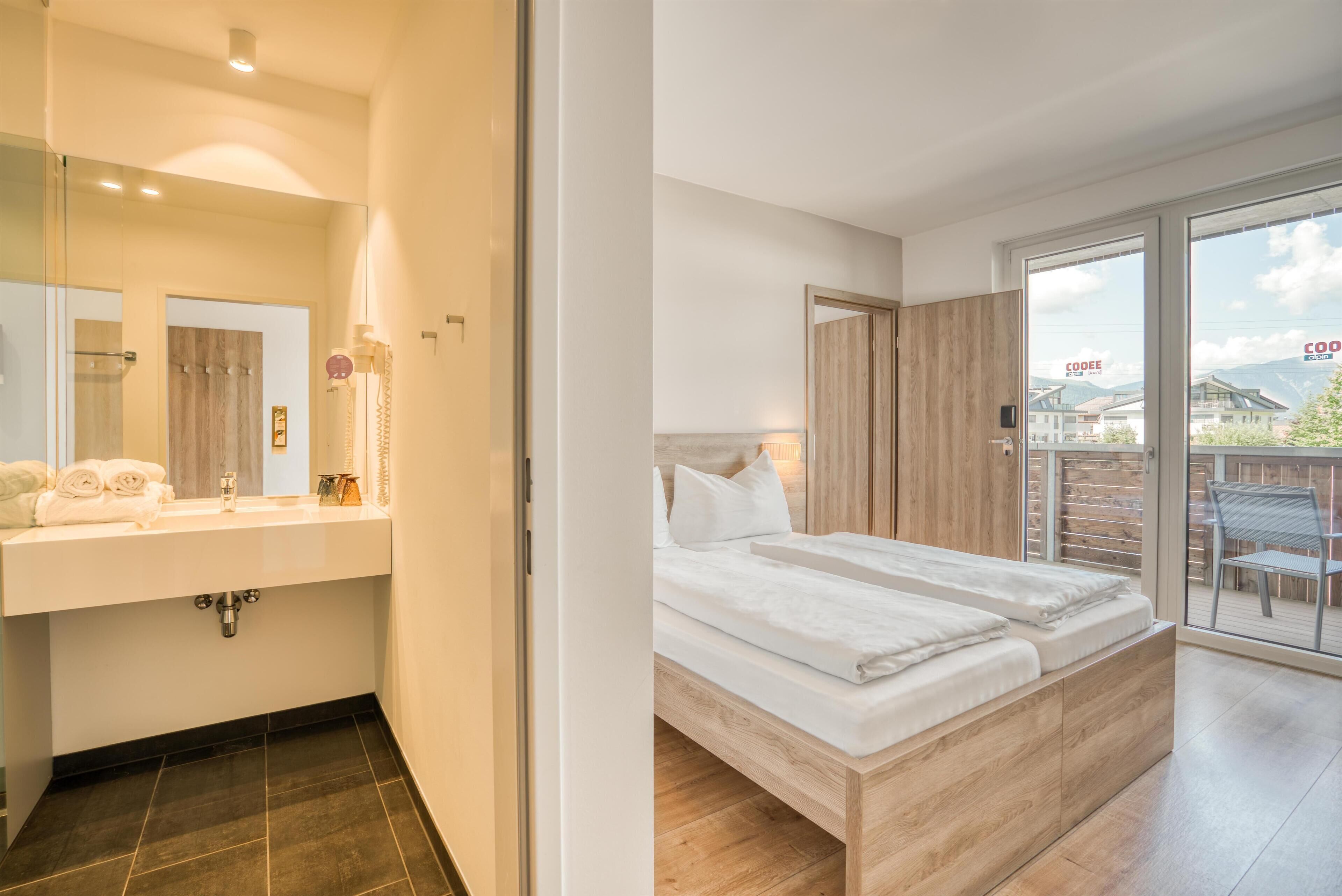 Un baño moderno con un espejo grande, un lavabo blanco y un suelo de baldosas oscuras. La habitación tiene una cama de madera con ropa de cama blanca y vistas a un balcón con mobiliario exterior.