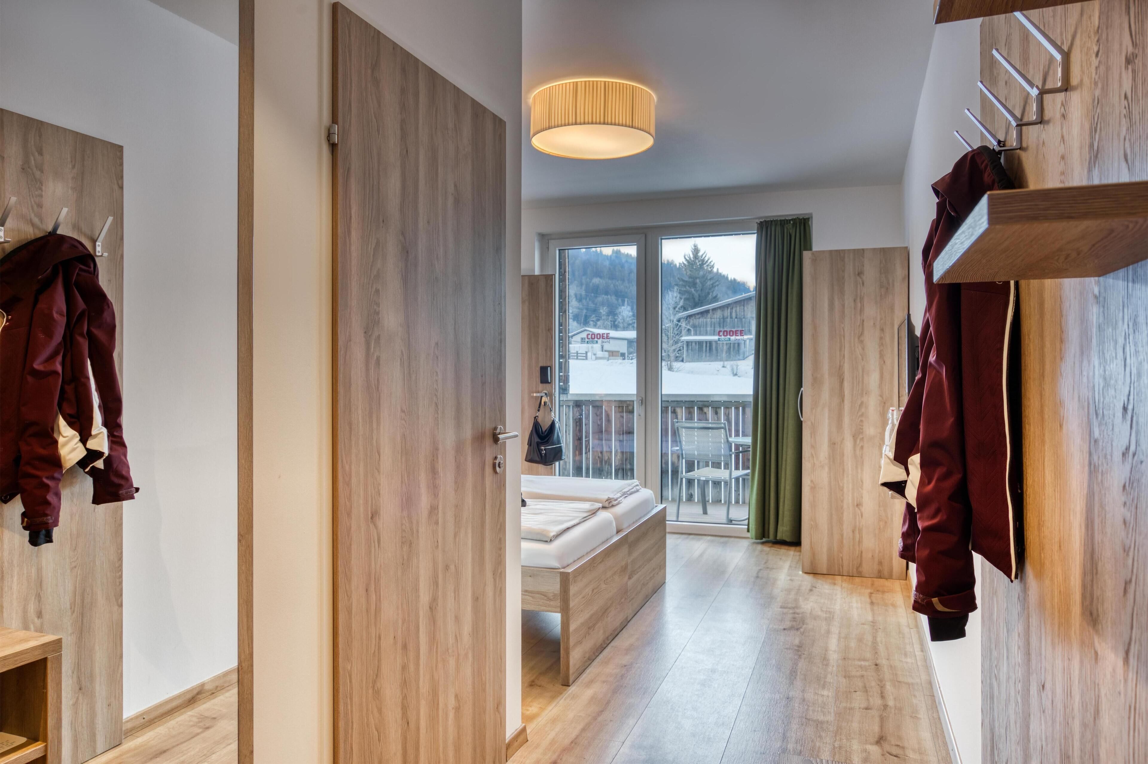 Une chambre d’hôtel moderne avec un plancher en bois, un lit avec un cadre en bois, une grande fenêtre donnant sur un paysage enneigé et un porte-manteau suspendu.