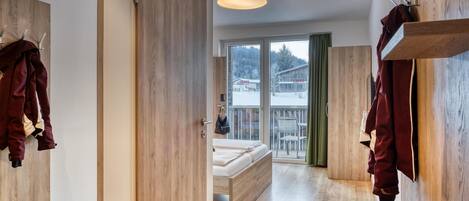 Une chambre d’hôtel moderne avec un plancher en bois, un lit avec un cadre en bois, une grande fenêtre donnant sur un paysage enneigé et un porte-manteau suspendu.