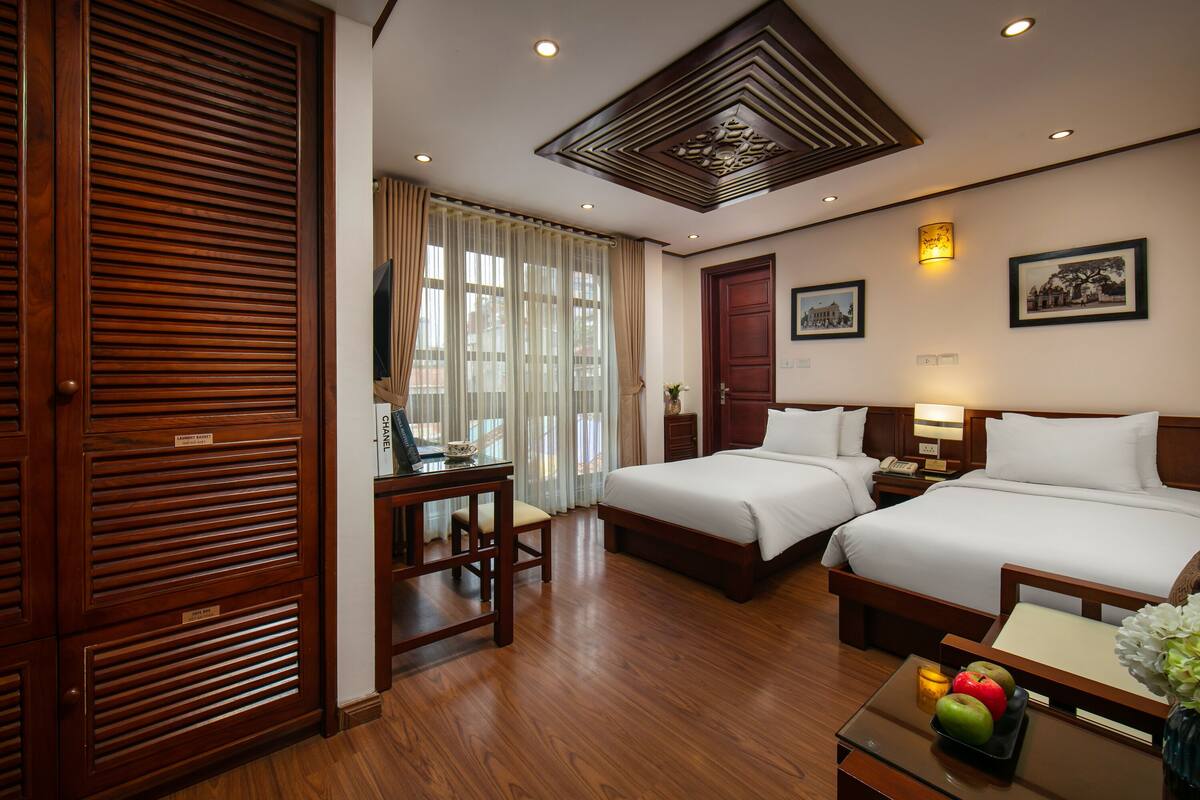 A hotel room with two beds, wooden flooring, a desk with a chair, a nightstand, and a table with fruit.