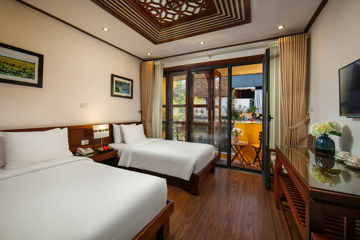 A hotel room with two beds, a wooden desk, a chair, a TV, and a balcony with a view of the outside.