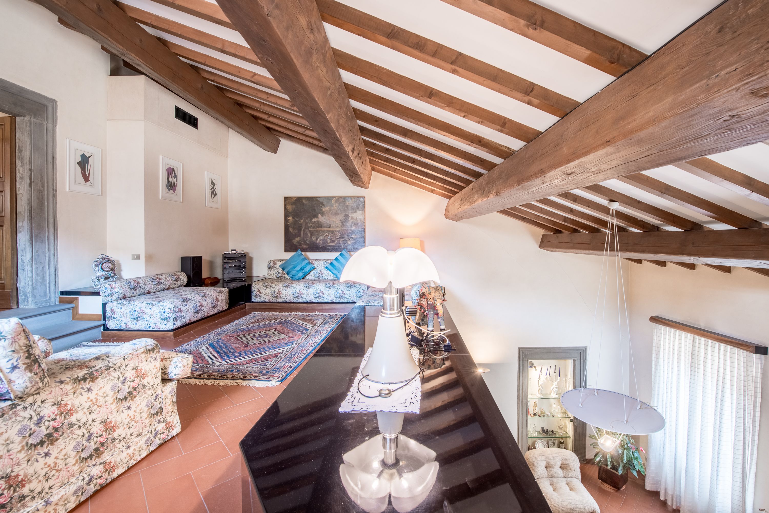 A spacious living room with a high ceiling, wooden beams, floral-patterned sofas, a central table, and a hanging light fixture.