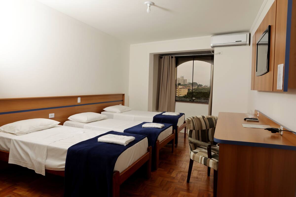 A hotel room with four single beds, a desk, a chair, and a window with a view of buildings.