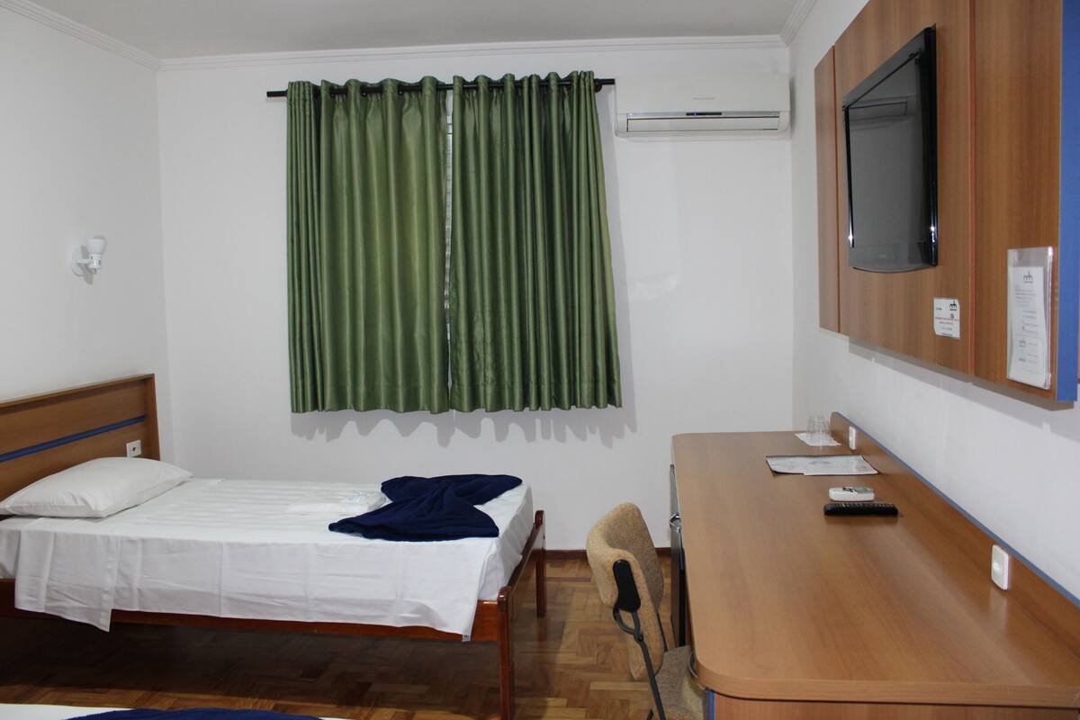 A hotel room with a bed, a desk, a chair, a TV, and a window with curtains.