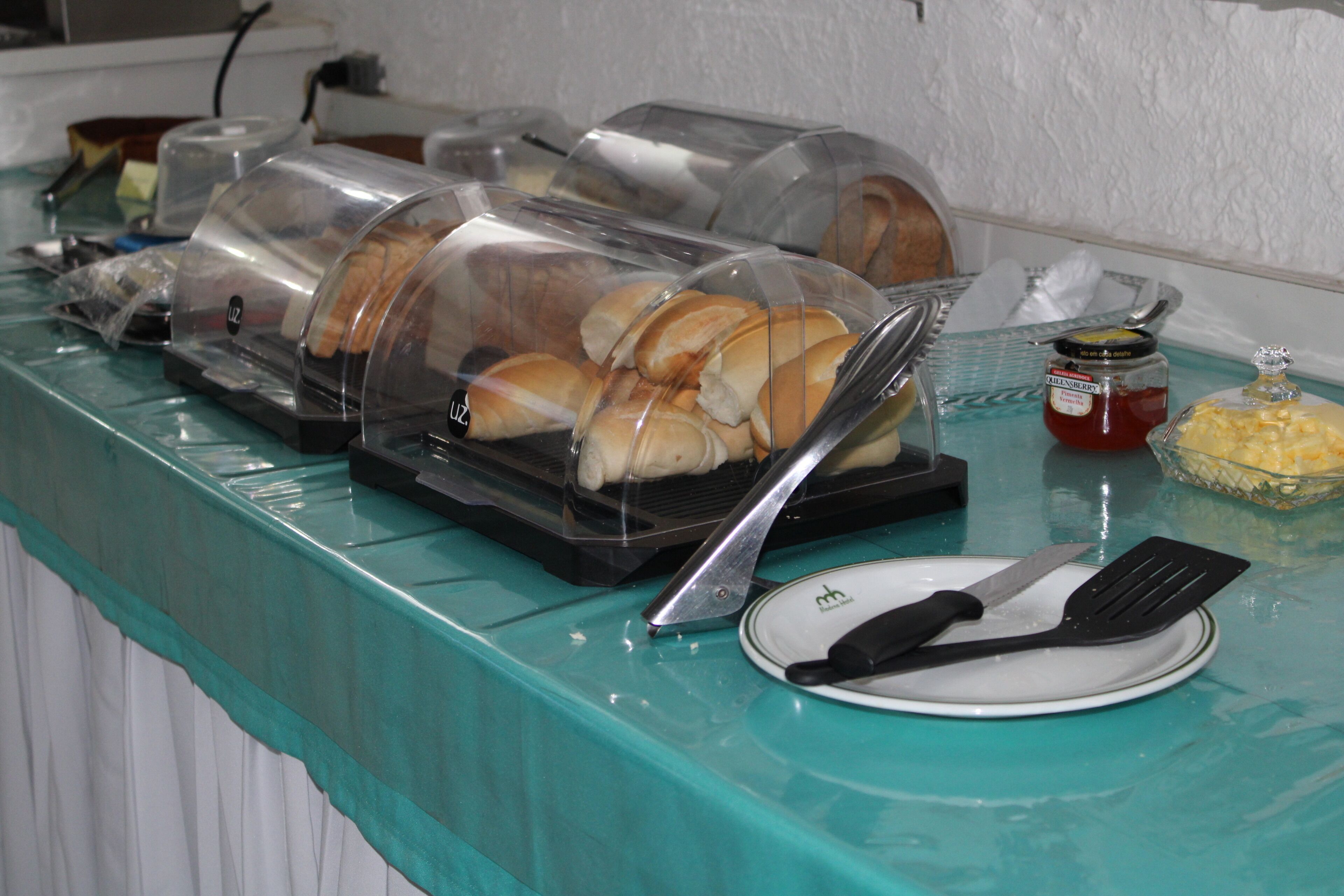 Disposição de buffet com distribuidores de alimentos cobertos, um prato com talheres e acompanhamentos em uma mesa azul-esverdeada.