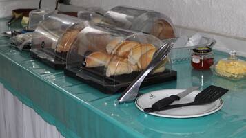 Disposição de buffet com distribuidores de alimentos cobertos, um prato com talheres e acompanhamentos em uma mesa azul-esverdeada.