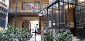 Un couloir étroit avec un balcon sur la gauche, une grande porte vitrée sur la droite et de la verdure au premier plan.