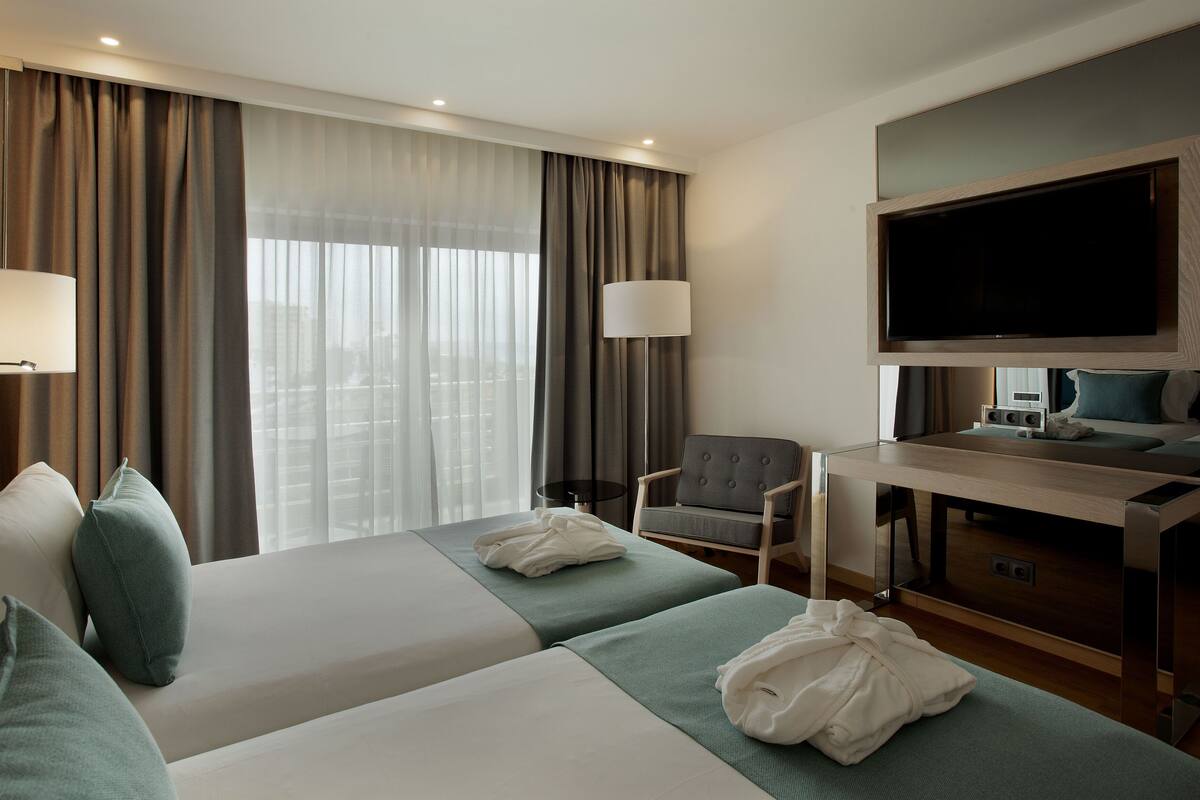 A hotel room with two beds, a desk, a chair, a TV, and a window with curtains.