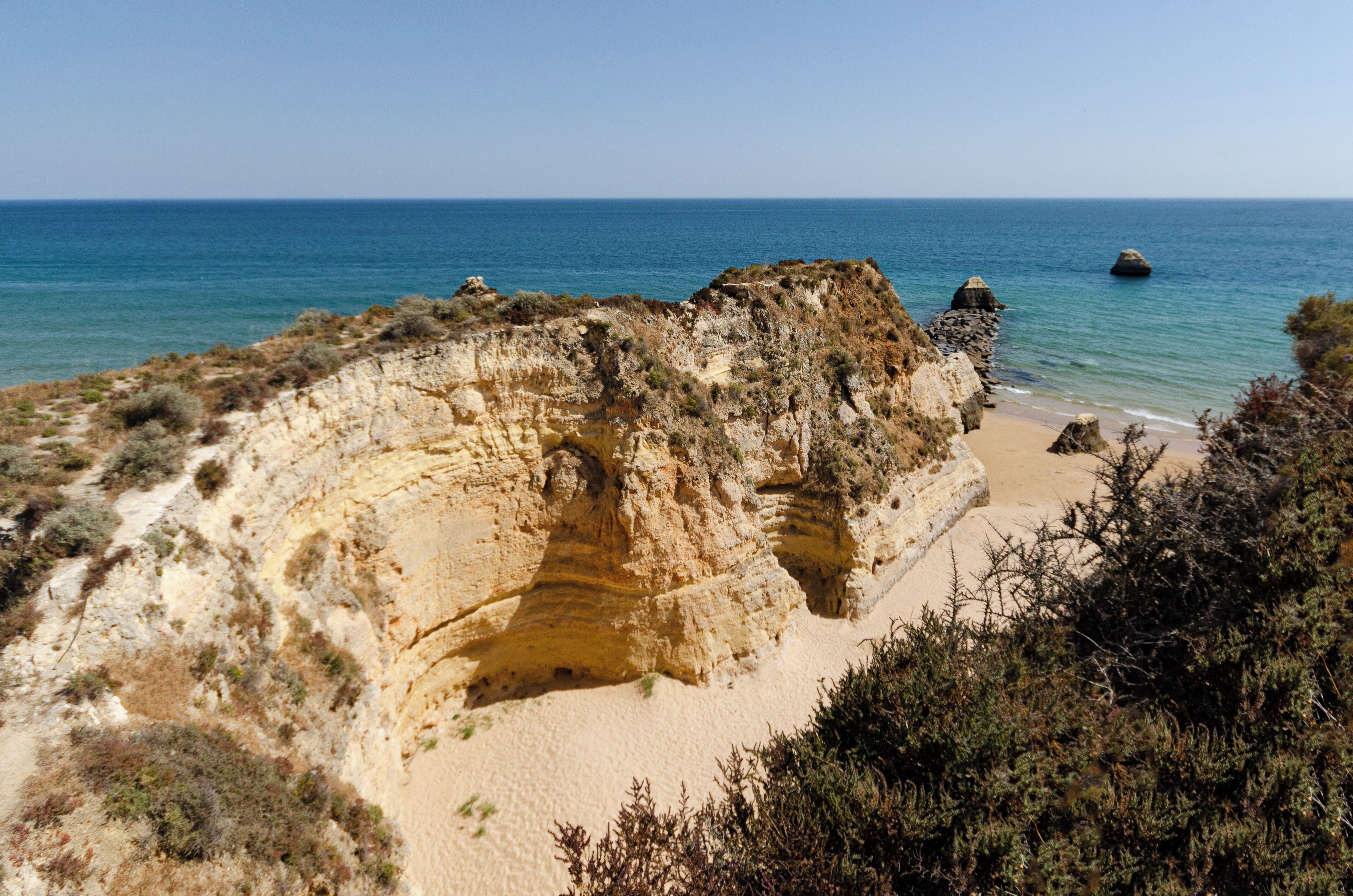 Paisaje costero con un acantilado imponente, una playa de arena y mar azul cristalino.