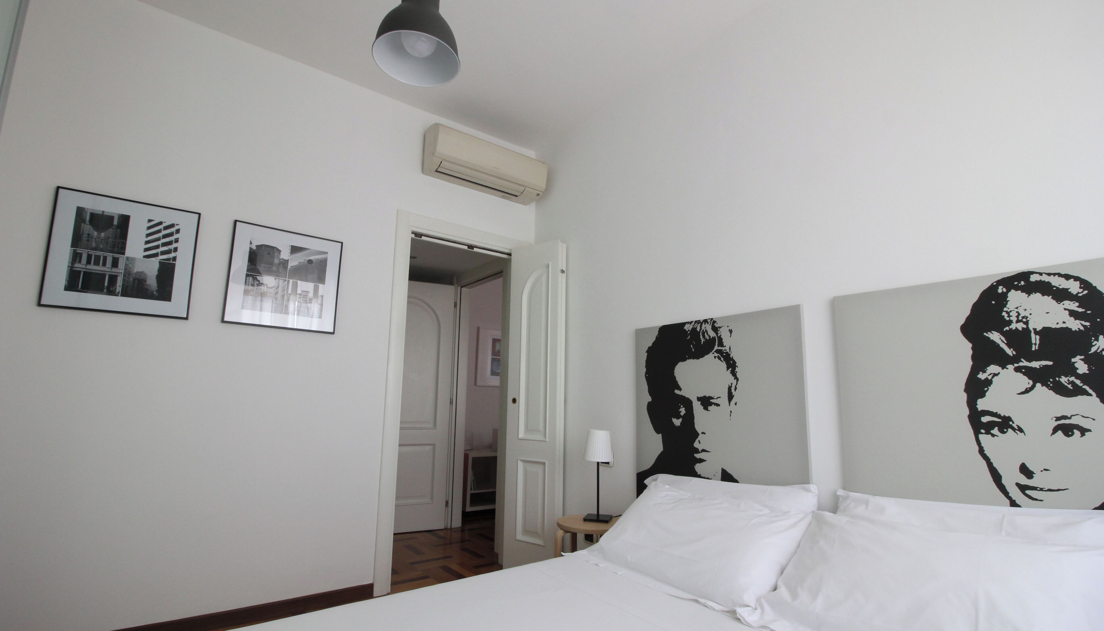 Hotellihuone, jossa on sänky, kaksi kehystettyä kuvaa, kattovalaisin ja ovi, joka johtaa käytävään.
