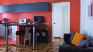 A modern kitchen with a microwave, stove, and dining area. There is a black sofa with a yellow pillow and a bookshelf. - Italianway   - Lodi 78 (Milan)
