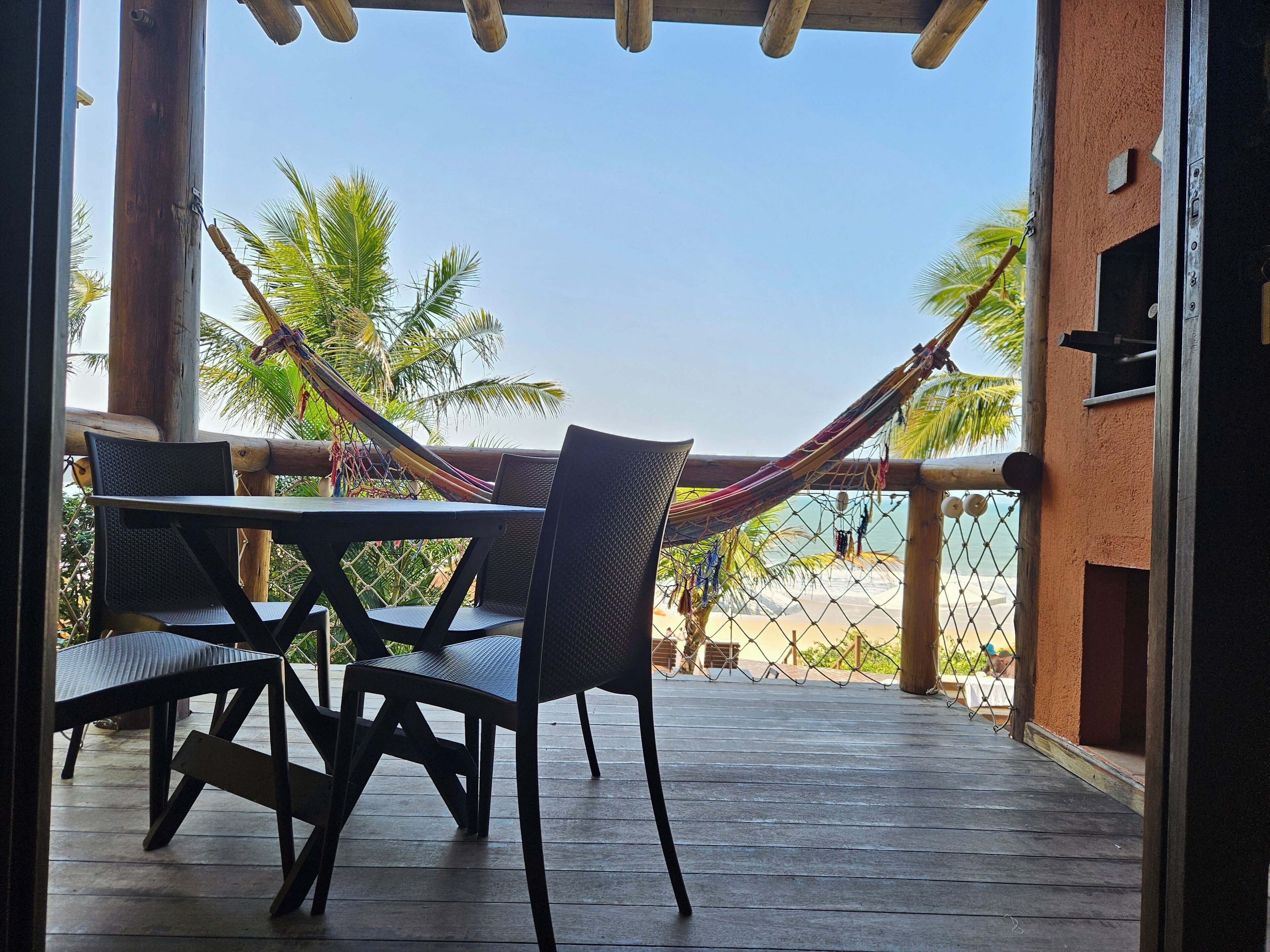 Una terraza de madera con una mesa, sillas, una hamaca y vistas a la playa y palmeras.
