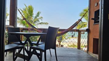 Una terraza de madera con una mesa, sillas, una hamaca y vistas a la playa y palmeras.