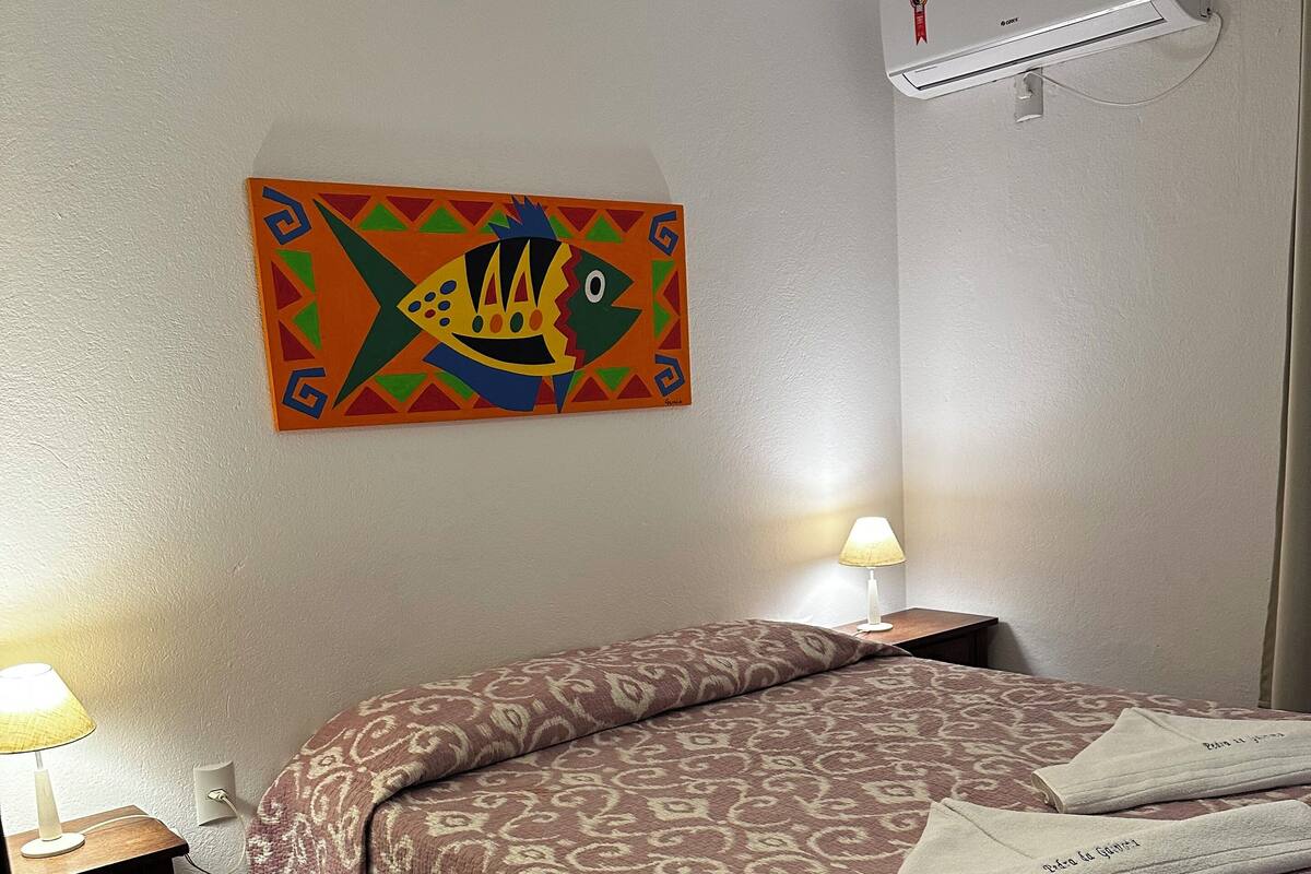 Ein Hotelzimmer mit Bett, Nachttisch mit Lampe, Klimaanlage und farbigem Wandbild.