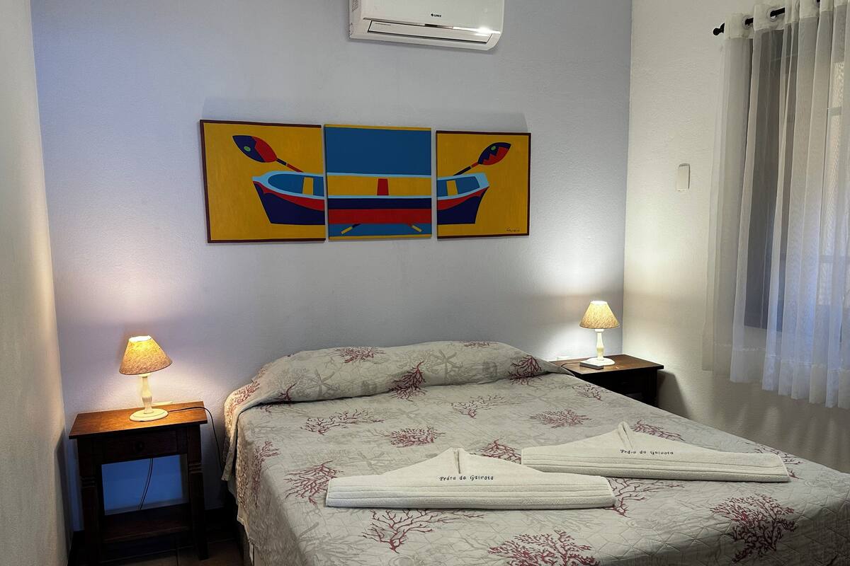 Ein Hotelzimmer mit einem Bett, zwei Nachttischen, einer Wand-Klimaanlage und einem Fenster mit durchsichtigen Vorhängen.