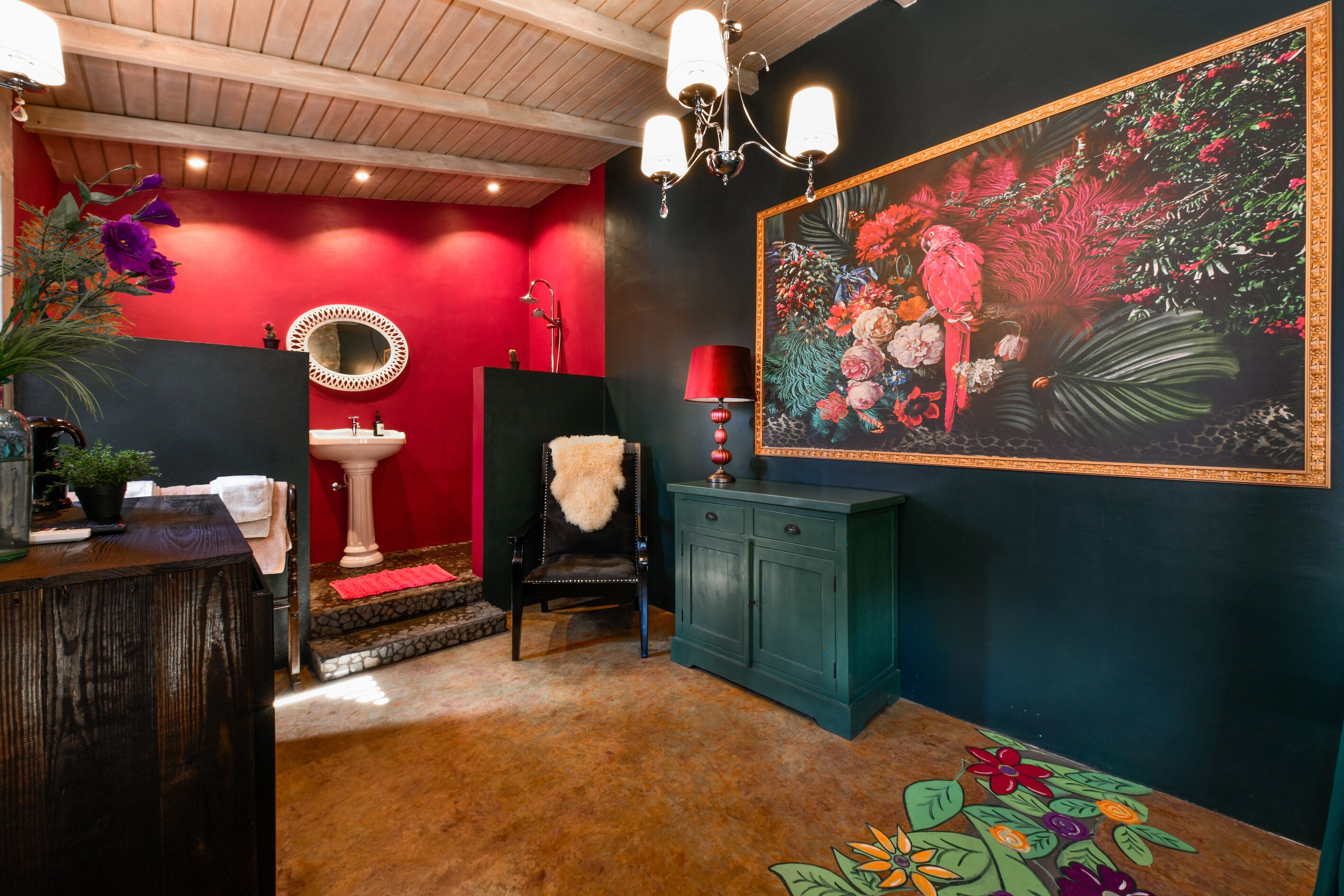 Une salle de bain avec un mur rouge, un lavabo sur pied blanc, une chaise noire recouverte d’un plaid en fausse fourrure, un meuble vert et un grand tableau représentant des motifs floraux.