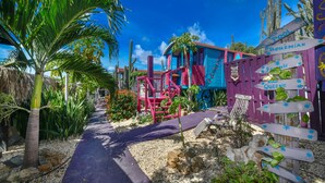 A colorful outdoor area with a pathway, a blue building, and a purple fence with directional signs. - Mammaloes (Santa Cruz)