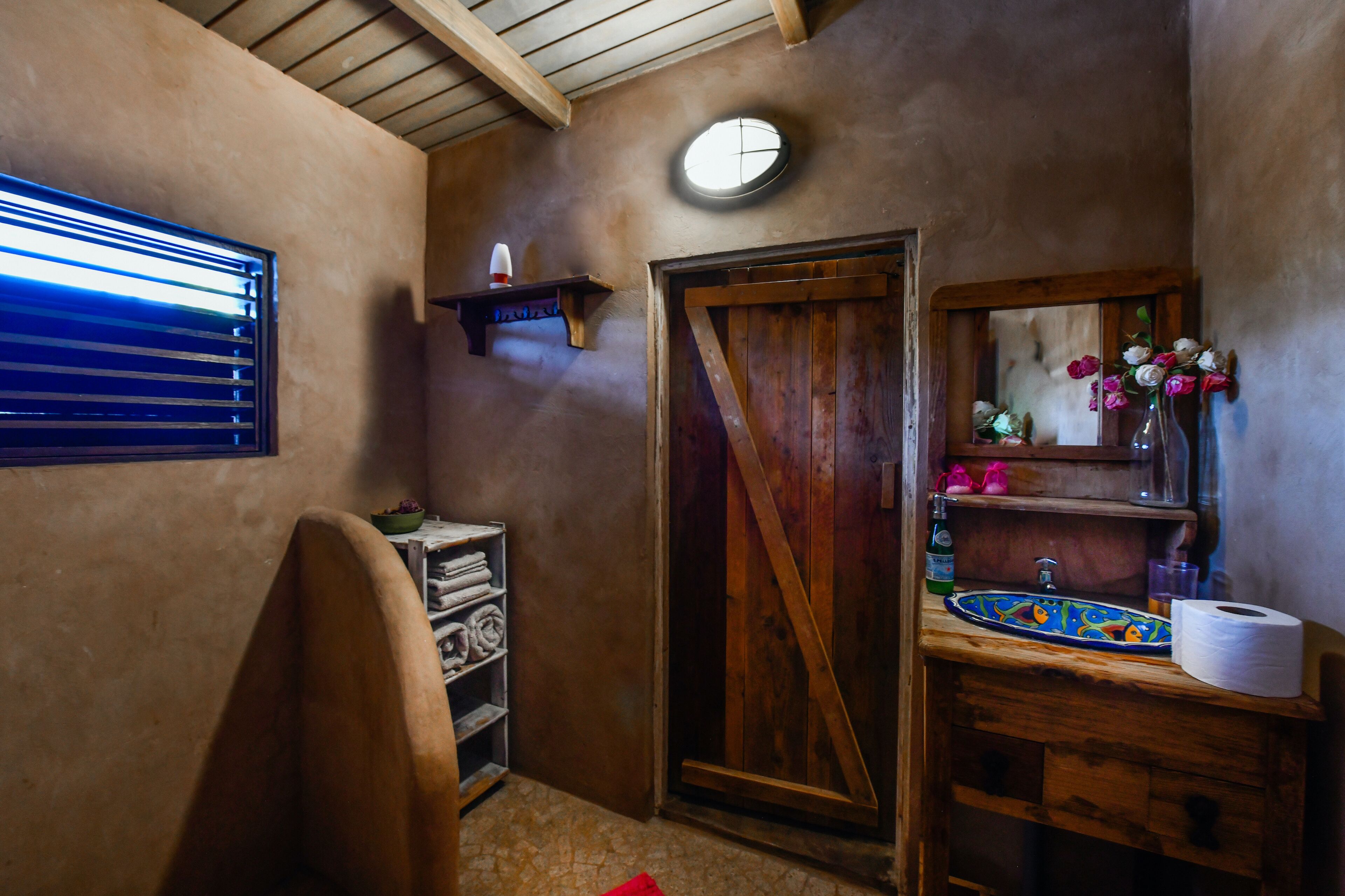 A rustic bathroom with a wooden door, a shelf with toiletries, and a mirror with a vase of flowers.
