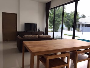 A modern living room with a wooden dining table, a dark brown sofa, and a flat-screen TV on a dark wooden stand.