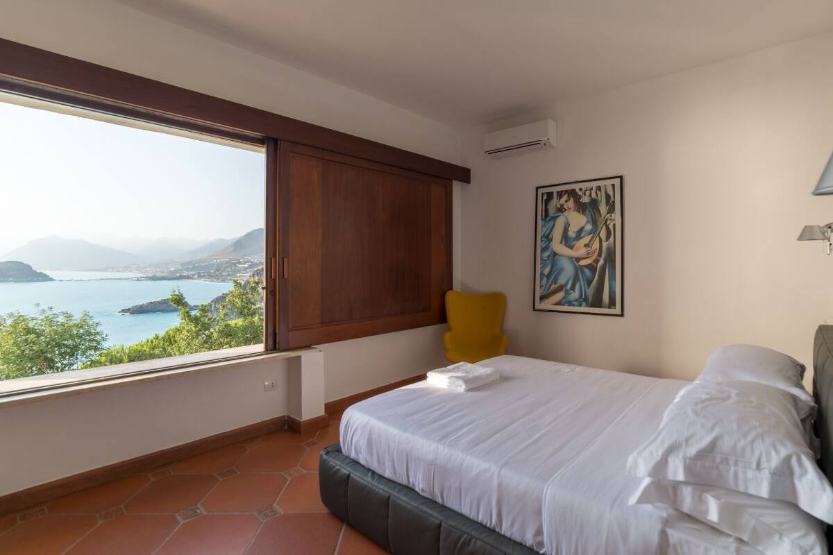 Quarto com cama, uma cadeira, um quadro e vista para o mar.
