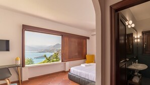 A bedroom with a large window overlooking a scenic view, a bed, a chair, a desk, and a bathroom with a sink and mirror. - Villa Crawford (San Nicola Arcella)